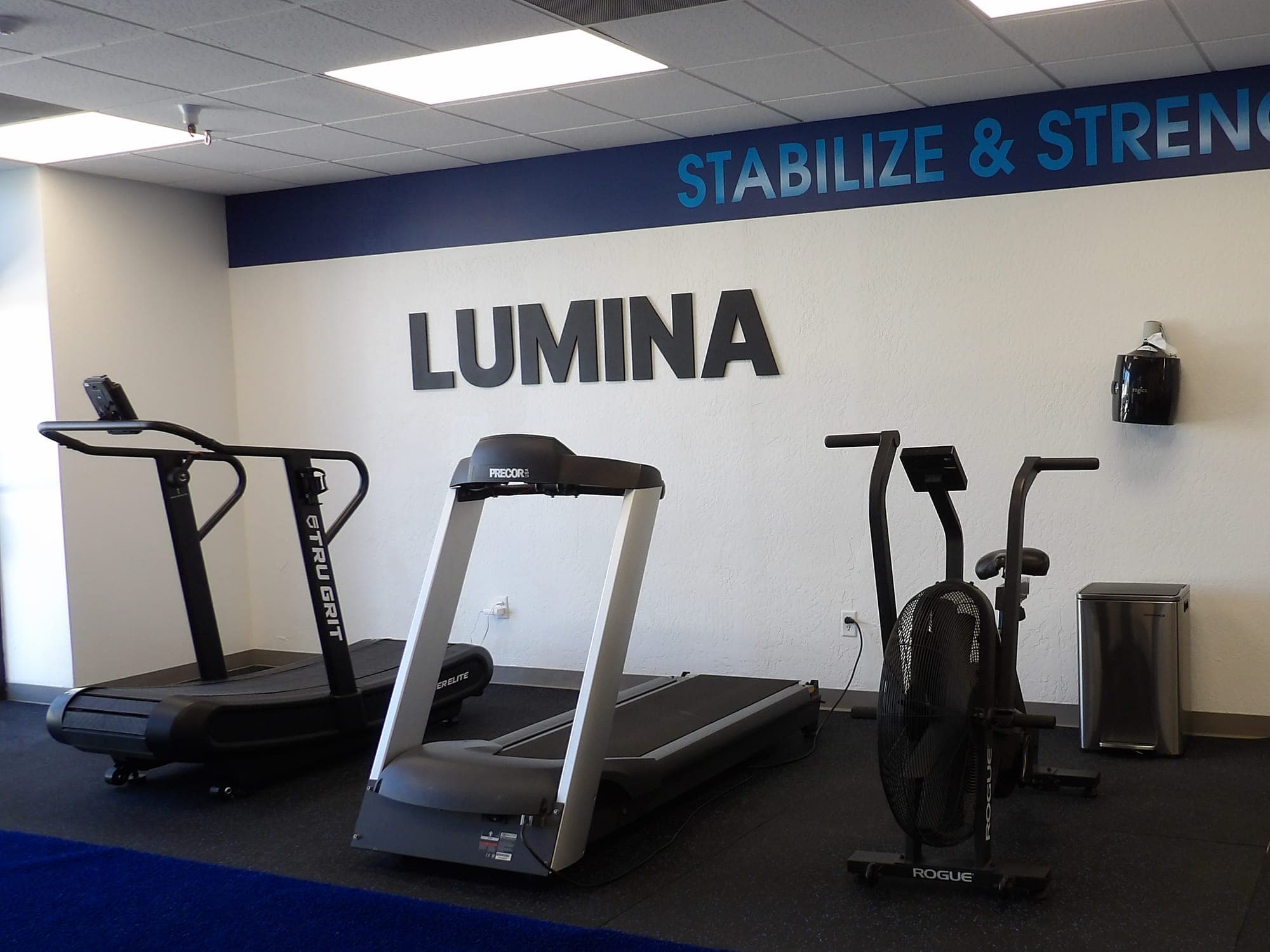 Cardio area at LUMINA with a curved manual treadmill, a motorized treadmill and an air bike below the LUMINA sign
